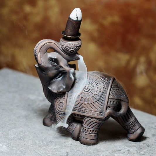 Ceramic elephant statue acts as an incense burner; smoke flows from trunk and top, set on a gray surface, with a blurred brown background.