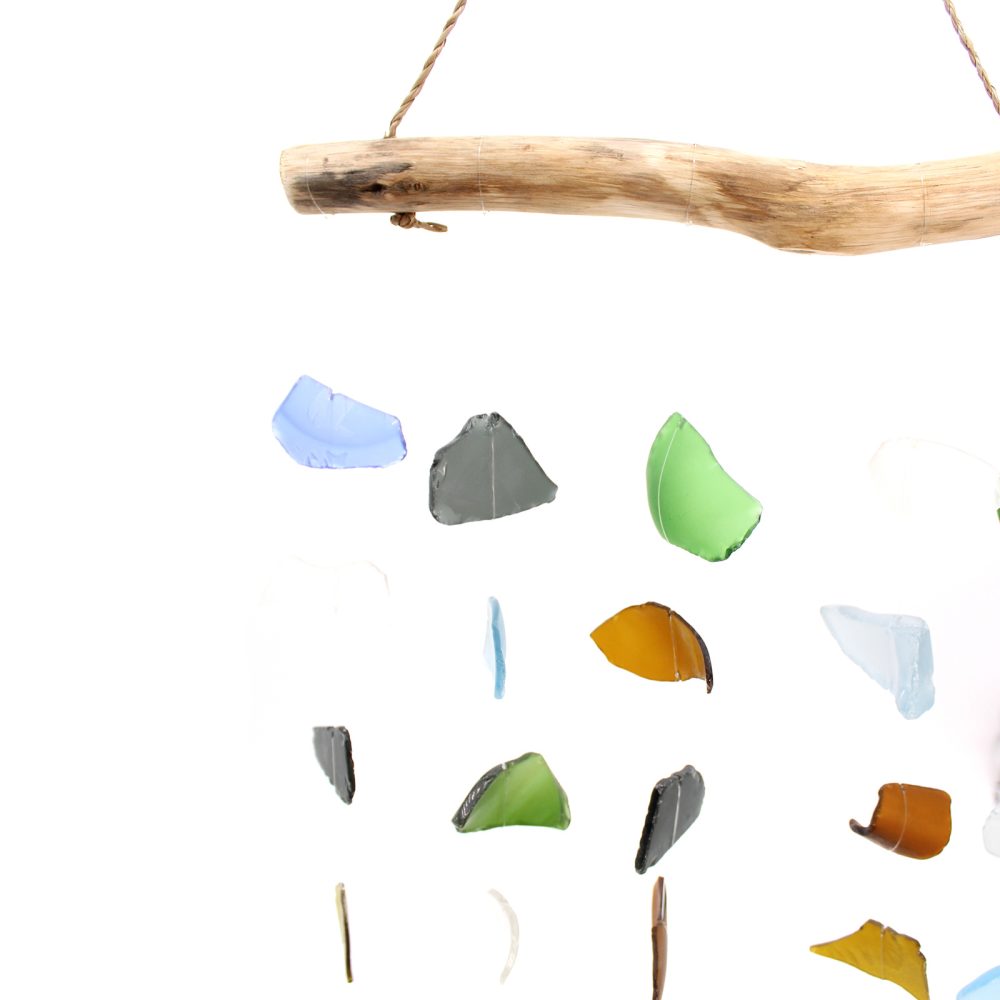 A driftwood branch hangs from twine, supporting a mobile of multicolored sea glass pieces. They dangle and move freely against a white background.