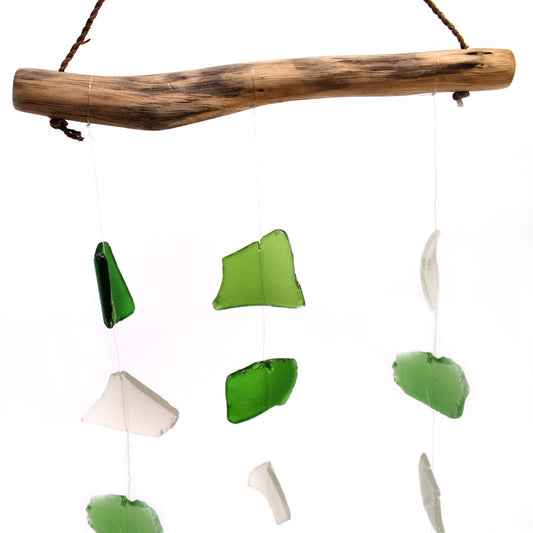A driftwood branch hangs from a string, suspending various pieces of green and white sea glass. The glass pieces dangle from thin threads against a white background.