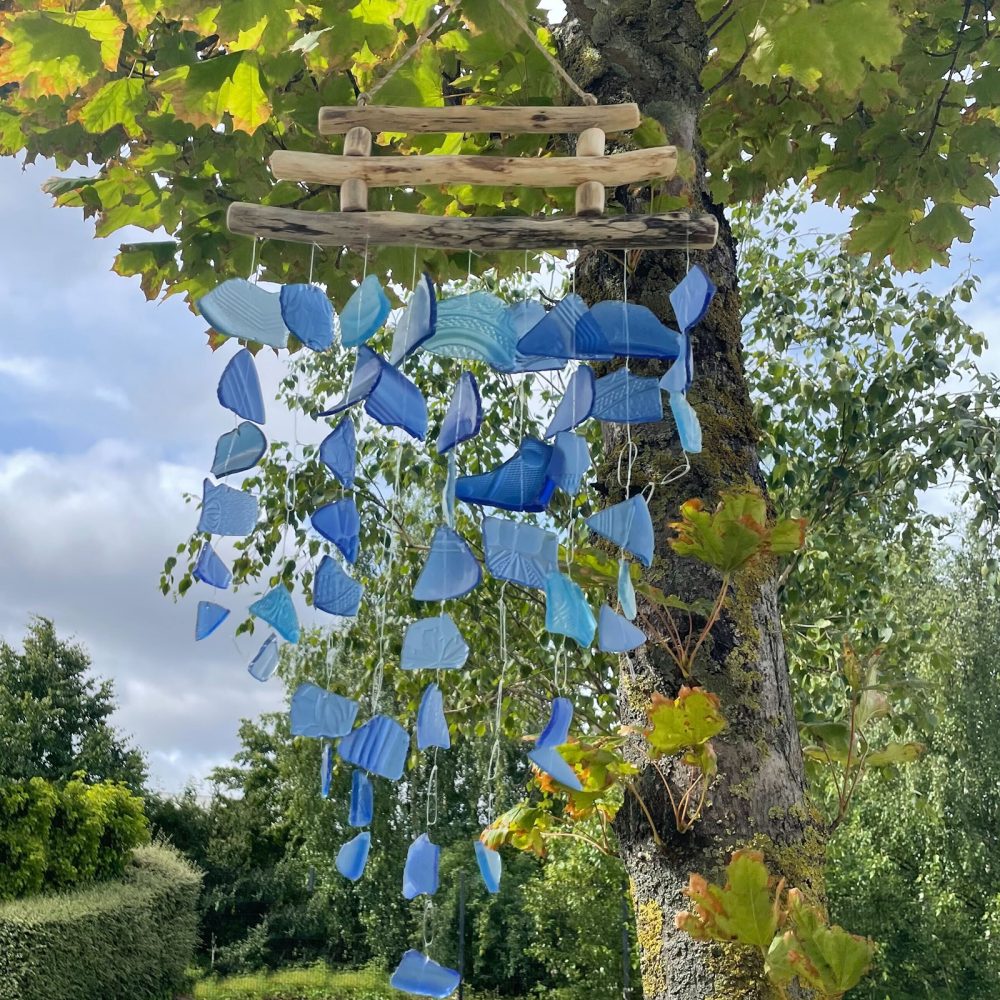 A driftwood mobile, adorned with various shades of blue glass pieces, hangs from a tree branch in a garden setting.