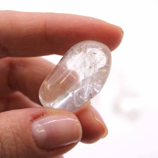 A clear, oblong crystal is being held between a thumb and index finger. The backdrop is a plain white surface.