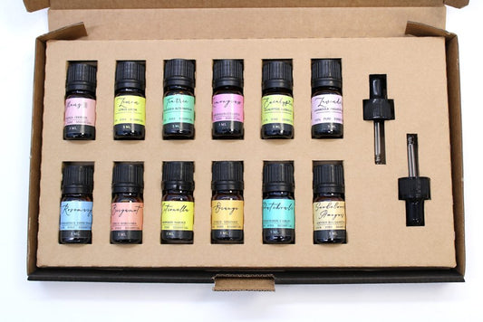 A cardboard box displays twelve small, amber bottles, each labeled with a different essential oil and species. Labels include "Ylang III," "Lemon," "Tea tree," "Lemongrass," "Eucalyptus Globulus," "Lavender," "Rosemary," "Bergamot," "Citronella," "Orange," "Patchouli," and "Sandalwood Amyris." Two black dropper tops are shown.