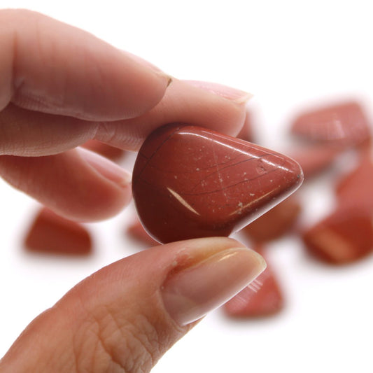 A red jasper gemstone is held between two fingers, the thumbnail of the lower finger visible. Other red jasper gemstones are visible, out of focus, on the white surface.