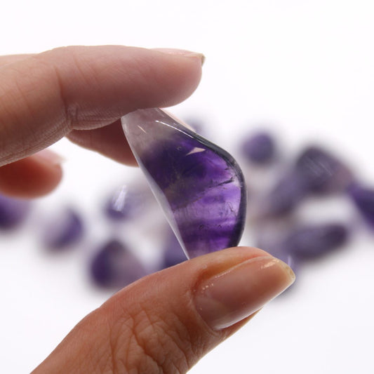 A hand holds a polished, purple amethyst gemstone between a thumb and finger. More amethyst stones are blurred in the background. All are set against a white background.