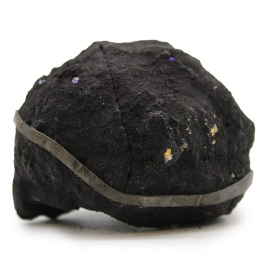 A rough, black rock speckled with purple and yellow rests on a white surface. A tarnished metal band encircles the rock's circumference.