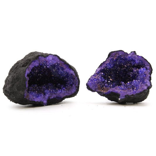 Two geode halves sit next to each other on a stark white surface. Both have jagged, dark gray exteriors and vibrant purple crystal-lined interiors.