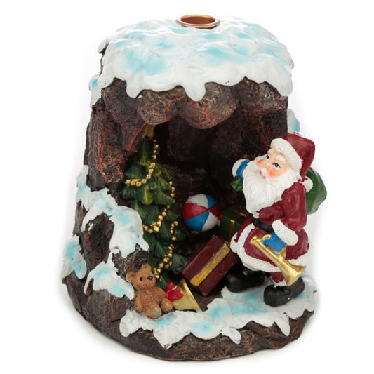 Santa figurine holding gifts and a trumpet stands beside a Christmas tree and teddy bear inside a snow-covered cavern.
