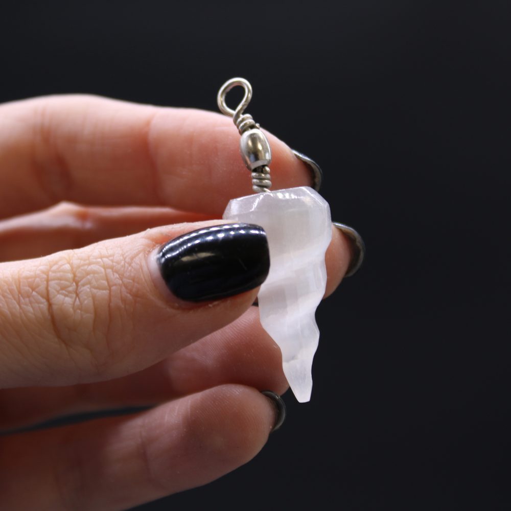 A hand with black nails holds a white, ridged crystal pendant with silver wire against a black background.