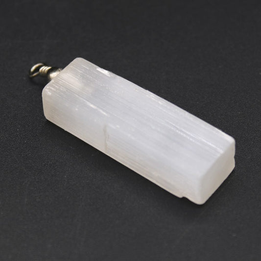 A selenite crystal pendant with a metal clasp rests diagonally against a stark black background. The translucent crystal is rectangular with subtle vertical ridges.