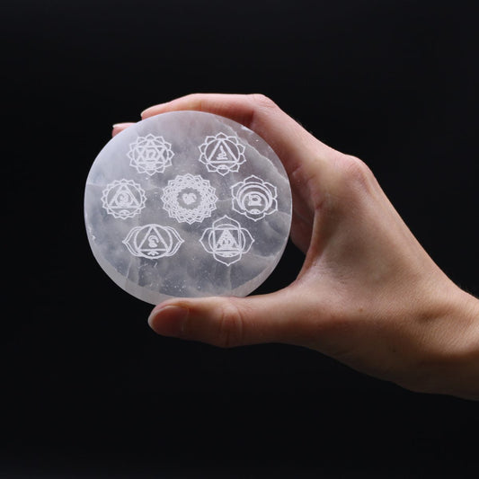 A hand holds a round selenite plate etched with seven chakra symbols. The background is black.