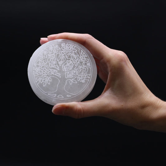A hand holds a round, white, flat stone etched with a "Tree of Life" design, against a black background.