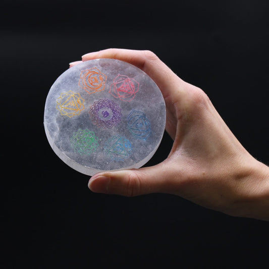 A hand holds a round, translucent stone disc against a black background. Seven colorful chakra symbols are etched onto the stone.