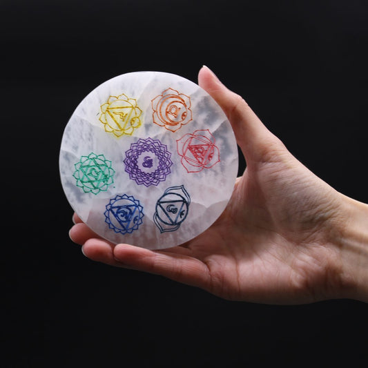 A round, white stone, etched with seven colorful chakra symbols, rests on a person's open palm against a solid black background.