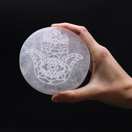 A hand holds a round, white selenite plate etched with a Hamsa hand design. The background is solid black.