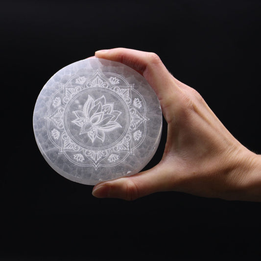 A hand holds a round, light-colored stone disc, etched with a lotus flower design, against a dark, solid background.
