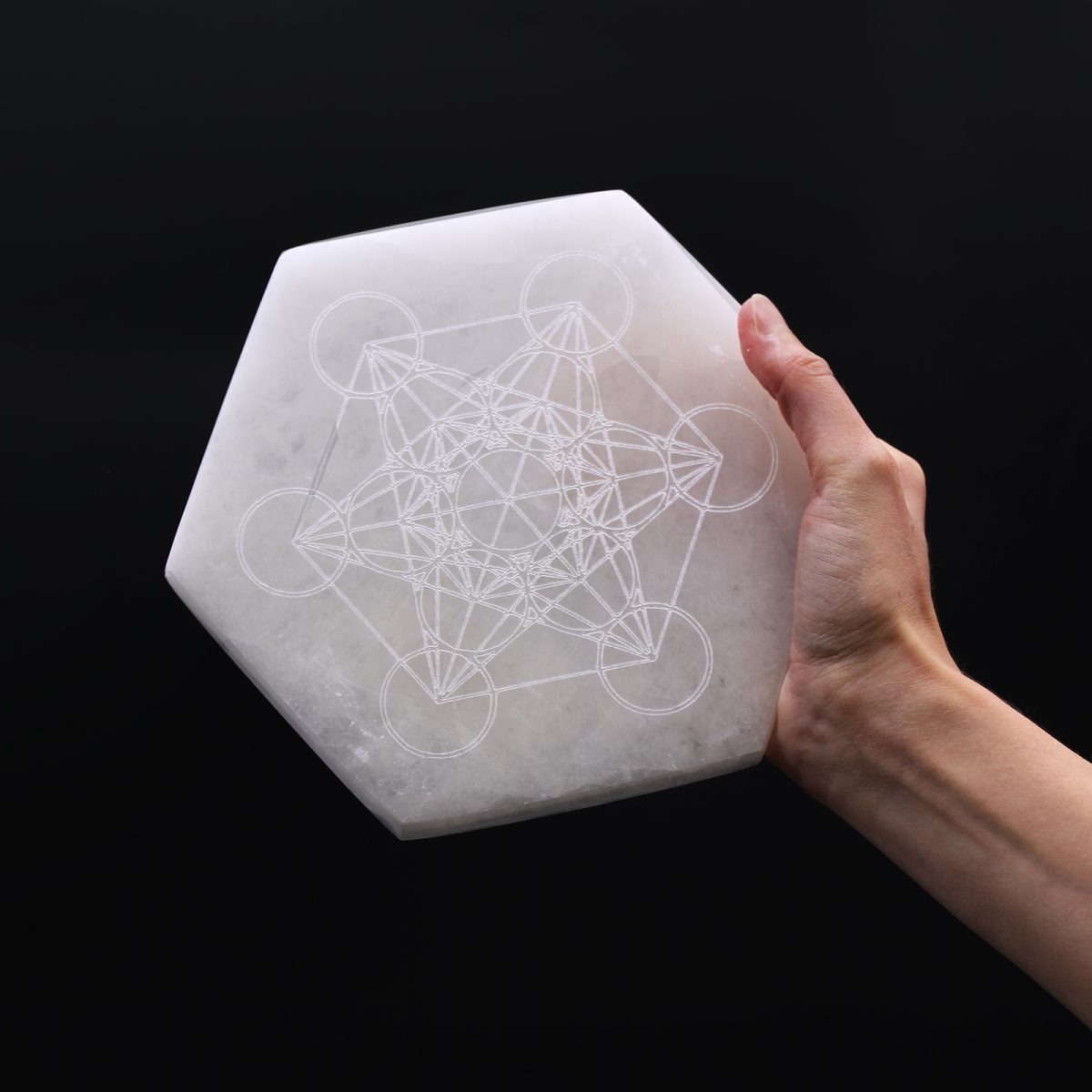 A hand holds a hexagonal selenite plate, engraved with a complex geometric pattern of circles and lines, against a solid black background.