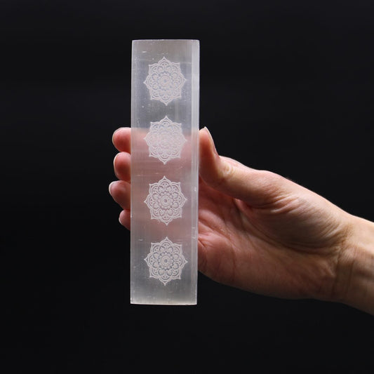 A hand holds a milky white, rectangular crystal. The crystal has four etched floral patterns, equally spaced. The background is solid black.