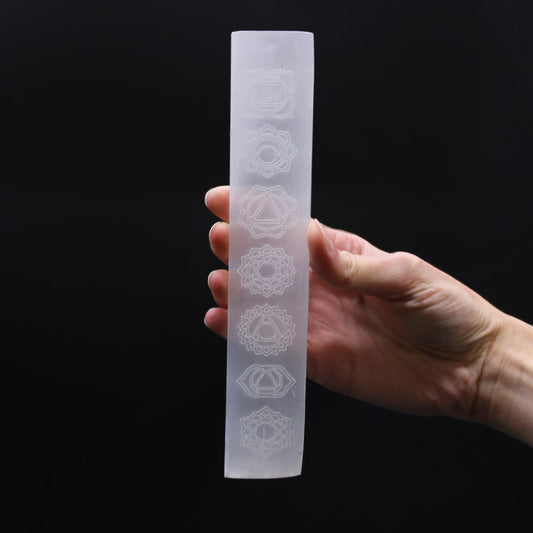 A hand holds a selenite rod, featuring etched chakra symbols. The background is a solid, dark color.
