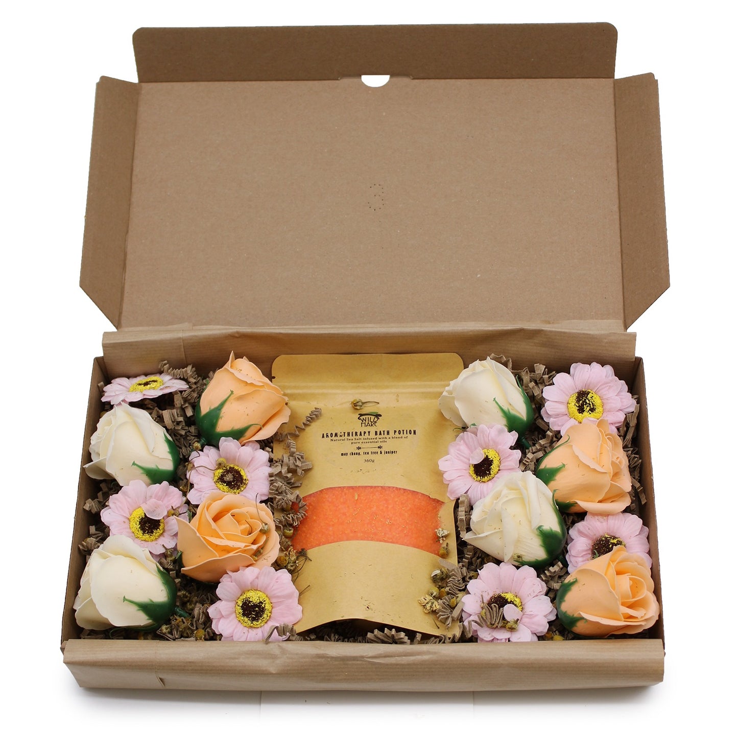 A brown cardboard box holds a gift set; a bag of orange bath salts sits centrally, surrounded by artificial roses and daisies. "WILD HARE AROMATHERAPY BATH POTION Natural Sea Salt infused with a blend of pure essential oils may chang, tea tree & juniper 360g" text is found on the bag.
