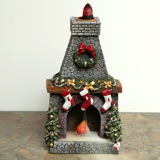 A small, decorative stone fireplace emits smoke, featuring holiday decorations with red stockings, a wreath, and bows. It sits on a stone-textured surface against a plain background.
