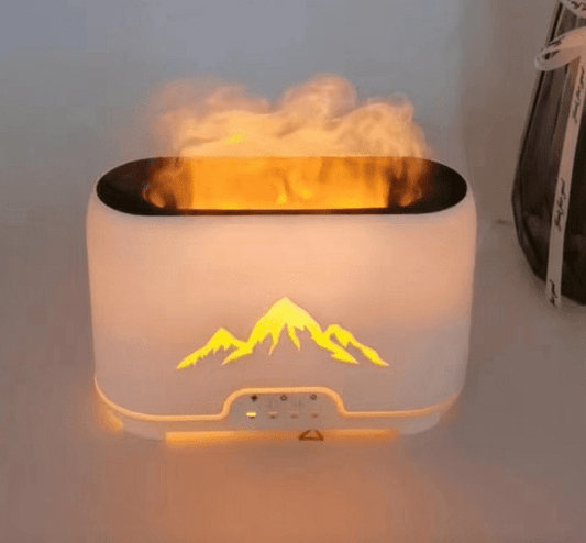 A white humidifier emits mist, illuminated by an orange glow, resembling flames. It features a mountain silhouette on the front. It sits on a table with a vase nearby.