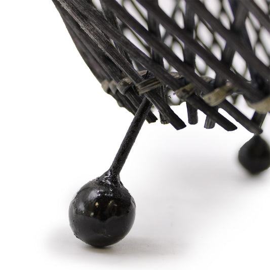 A black woven basket stands on a single, rounded leg. The basket is dark-stained and rests on a white surface.