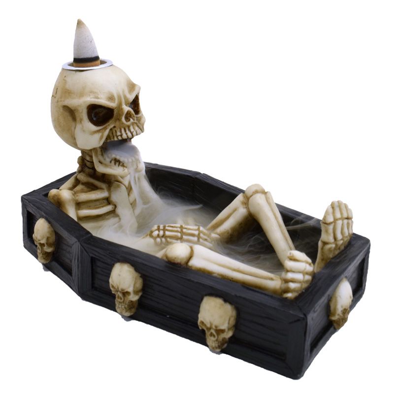 A skeleton reclines inside a black coffin decorated with skulls, exhaling smoke. Its head supports a cone-shaped object, suggesting incense use, in a spooky atmosphere.