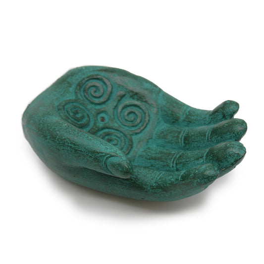 Sculpted turquoise hand with intricate spiral designs in the open palm, resting on a white surface, suggesting a decorative or symbolic object in a minimalist setting.