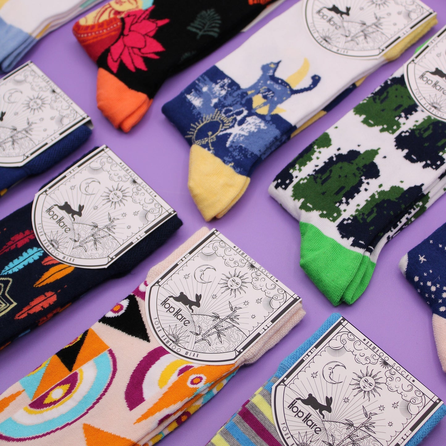 Colorful socks with various designs are arranged on a purple surface. Each pair is packaged with a tag featuring a rabbit, sun, moon, and bamboo. The tag reads "Hop Hare YOUR BAMBOO WEAR".