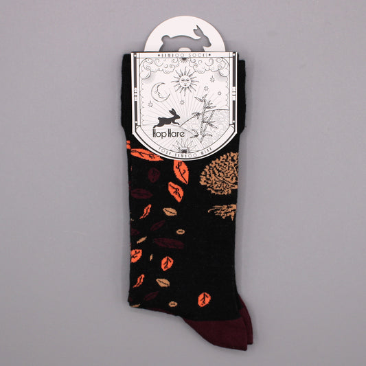 A pair of black socks, featuring an autumnal leaf pattern in orange and brown, hangs from a tag against a gray background. The tag reads "*BAMBOO SOCKS*", depicts a rabbit, sun, moon, and bamboo, and states "Hop Hare YOUR BAMBOO WEAR".