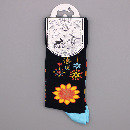 A pair of dark-colored socks, adorned with colorful floral designs, hangs from its packaging against a gray backdrop. The packaging displays the brand name "Hop Hare" and describes the socks as "Bamboo Socks."
