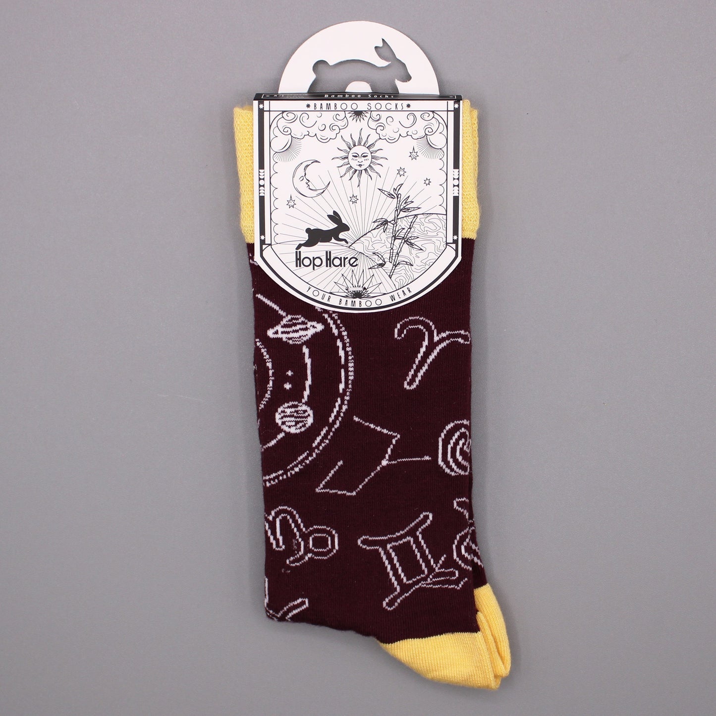 A maroon sock with a yellow cuff, featuring white zodiac constellations, hangs from its packaging against a gray background. The packaging displays: "*BAMBOO SOCKS*", a rabbit, sun, moon, bamboo, and "Hop Hare YOUR BAMBOO WEAR".