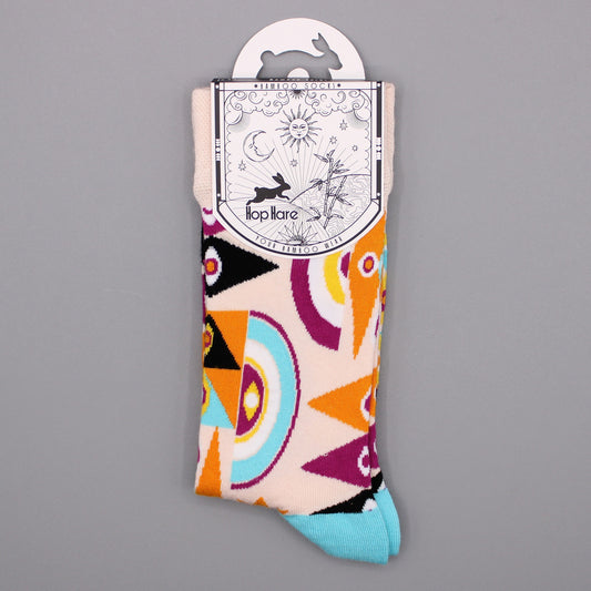A pair of socks, displayed on a gray background, features a geometric pattern in orange, purple, teal, and black on a cream base. The socks' packaging shows a rabbit, sun, moon, and bamboo. "Hop Hare YOUR BAMBOO WEAR" is printed on the tag.