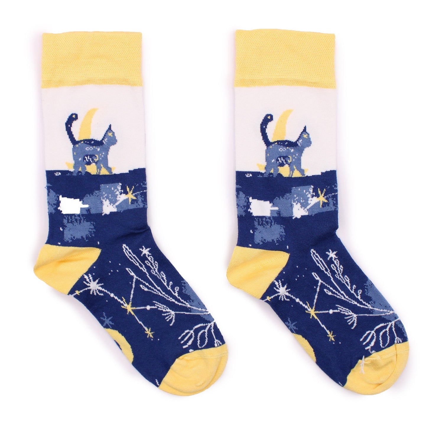 A pair of socks, displaying a cat sitting on a crescent moon amidst stars and constellations, are laid flat against a white background.