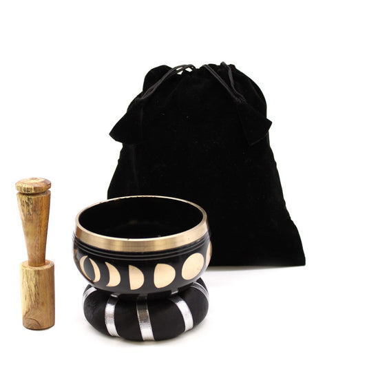 A Tibetan singing bowl with moon phases design rests on a black cushion with silver stripes. A wooden mallet stands beside it. A black drawstring bag is in the background.