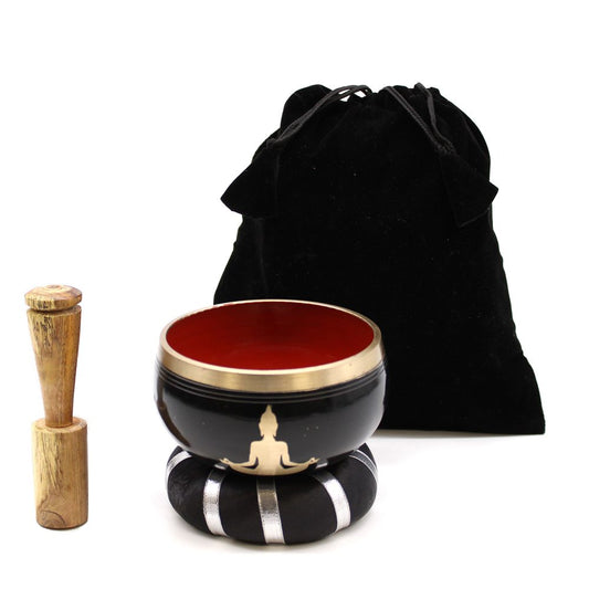 A black singing bowl sits on a striped cushion, with a wooden mallet beside it. A black drawstring bag is in the background. The bowl's interior is red, featuring a simple figure design.