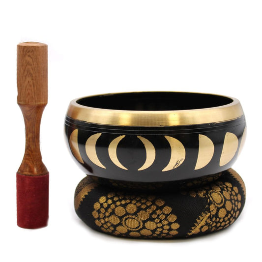 A black singing bowl with golden moon phase designs rests on a patterned cushion. A wooden mallet with a red tip is positioned beside it against a white background.