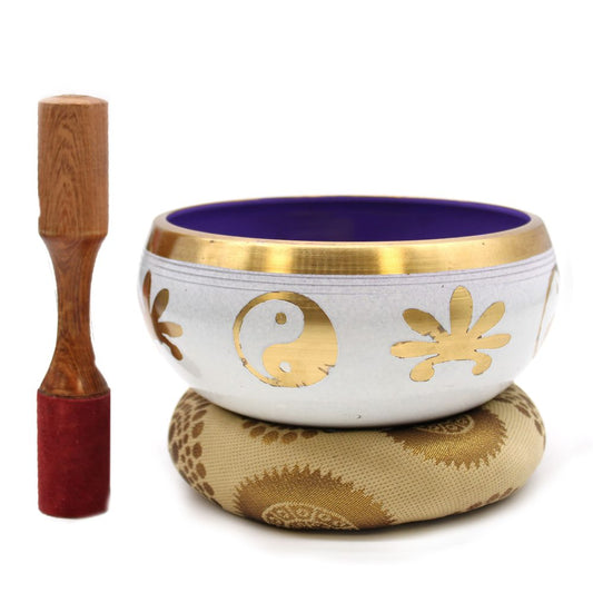A metallic singing bowl with gold yin-yang and floral symbols rests on a patterned cushion. A wooden mallet with a red felt tip stands upright beside it, set against a plain background.