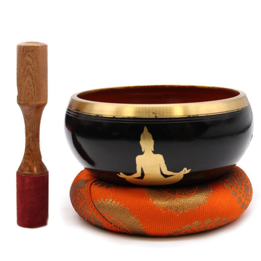 A black singing bowl with gold Buddha silhouette rests on an ornate orange cushion. A wooden mallet with a red suede handle stands upright beside it.