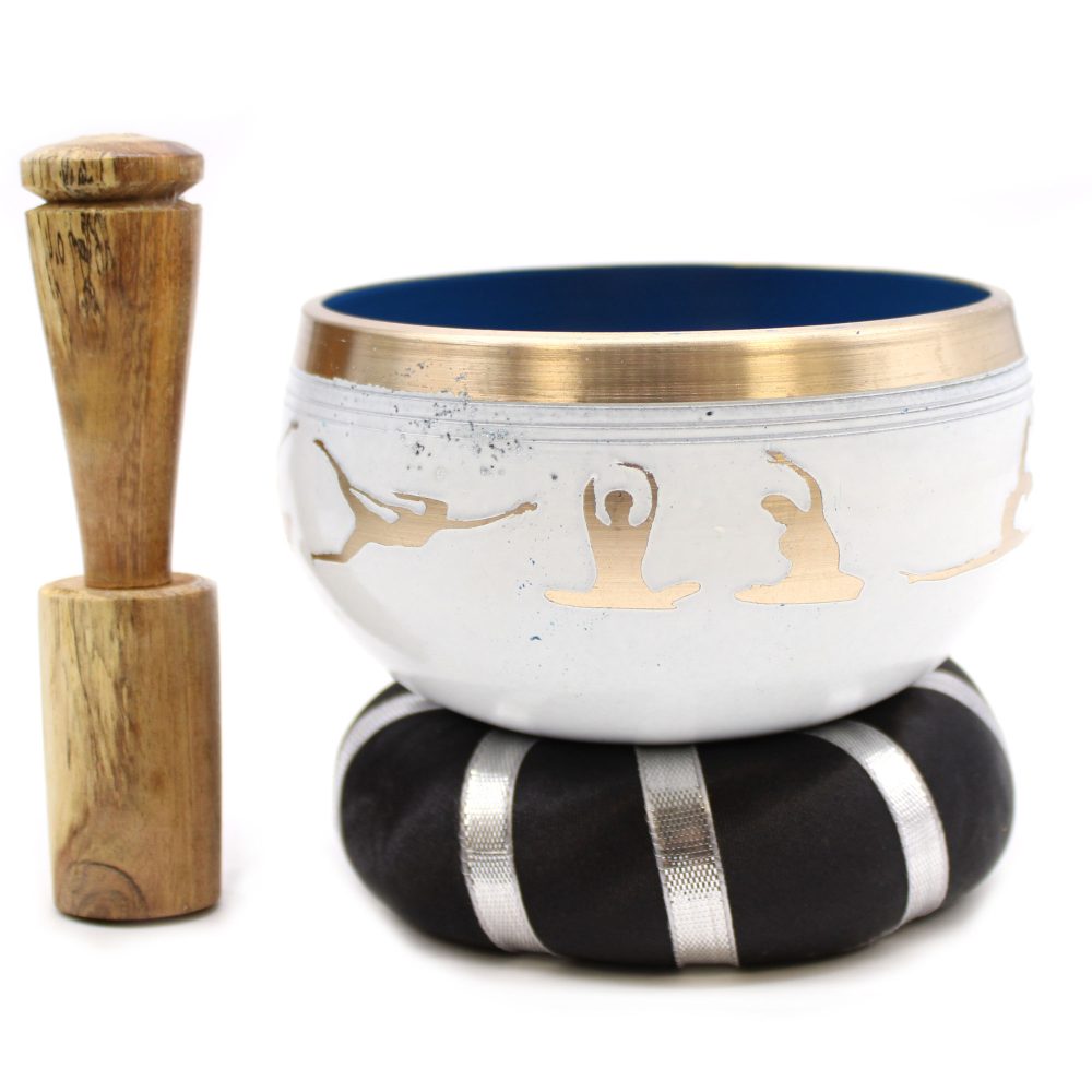 A silver singing bowl, decorated with gold yoga silhouettes, rests on a black cushion with silver stripes. Beside it, a wooden mallet stands upright, suggesting preparation for sound therapy.