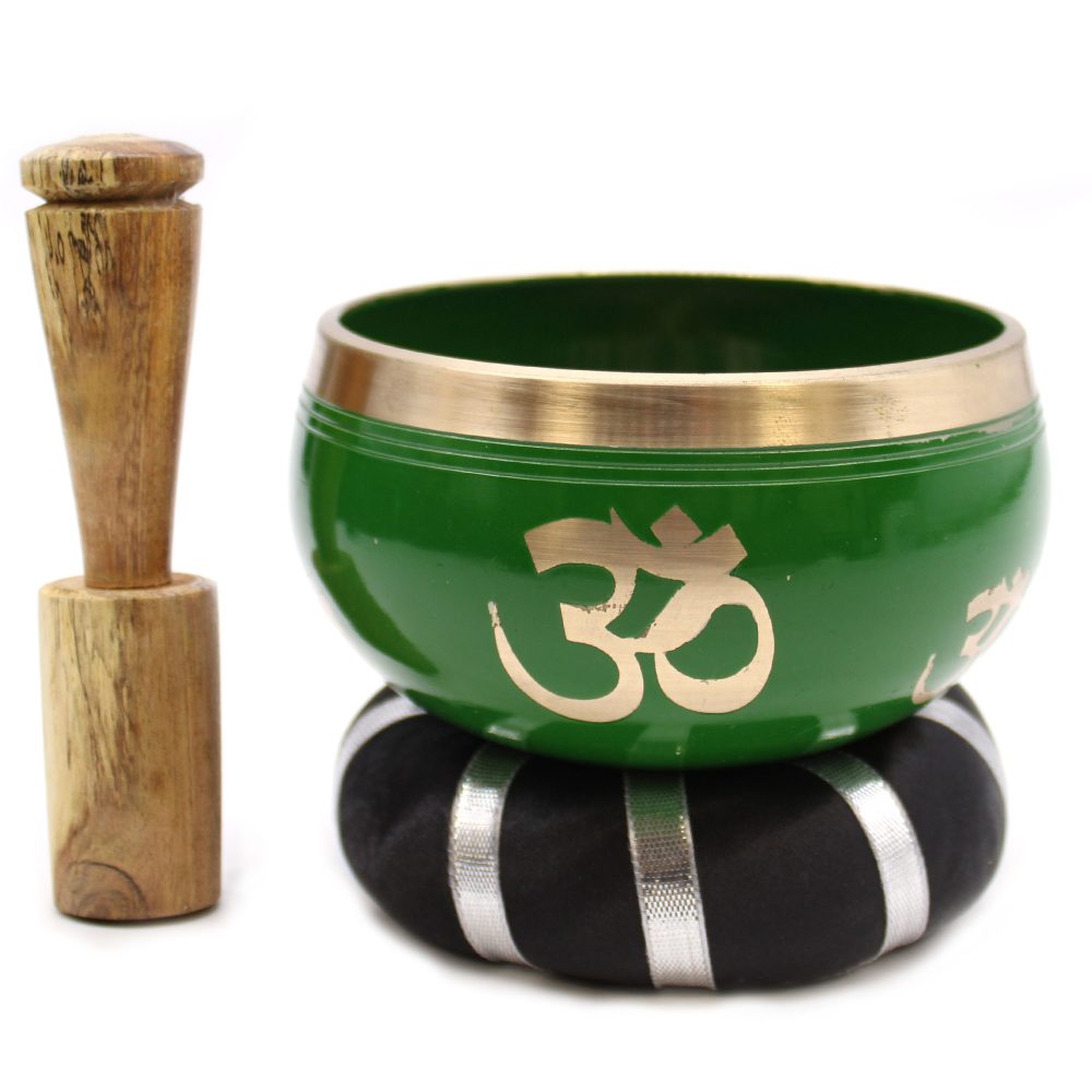A green singing bowl with golden "Om" symbols rests on a black cushion with silver stripes. A wooden mallet stands upright beside it.
