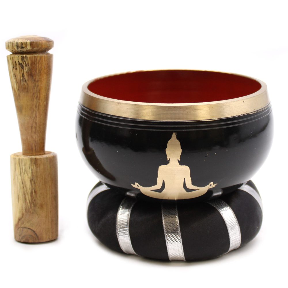 A black singing bowl with a golden meditating figure rests on a black cushion with silver stripes. A wooden striker stands upright beside it, set against a plain background.