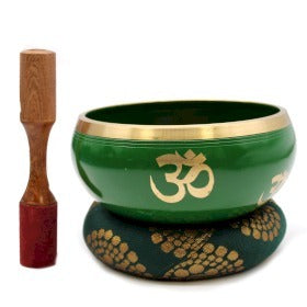 A green singing bowl with golden "Om" symbols rests on a patterned cushion. A wooden striker with a red tip stands beside it. The background is plain white.
