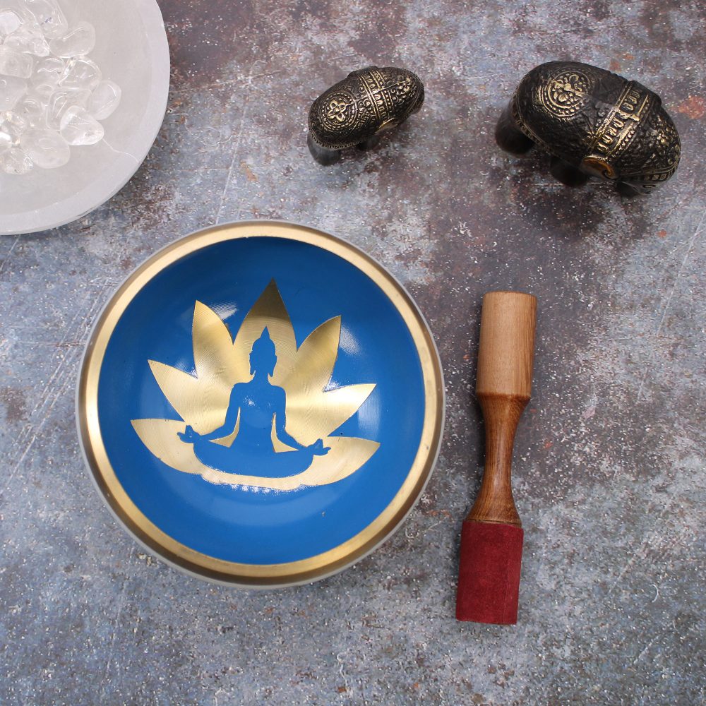 A blue singing bowl with a gold lotus and meditating figure is surrounded by decorative elephants, a wooden mallet, and a bowl of clear stones on a textured surface.
