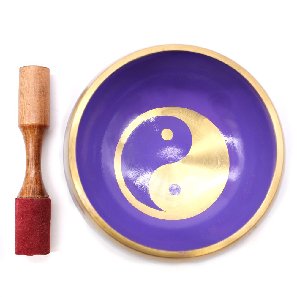 A purple singing bowl with a gold yin-yang symbol inside is placed next to a wooden mallet with a red suede tip, on a plain white surface.