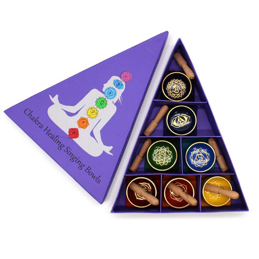 Purple triangular box displaying chakra healing singing bowls, each with a wooden striker. The lid shows a meditating figure with colored chakras. Text: "Chakra Healing Singing Bowls."