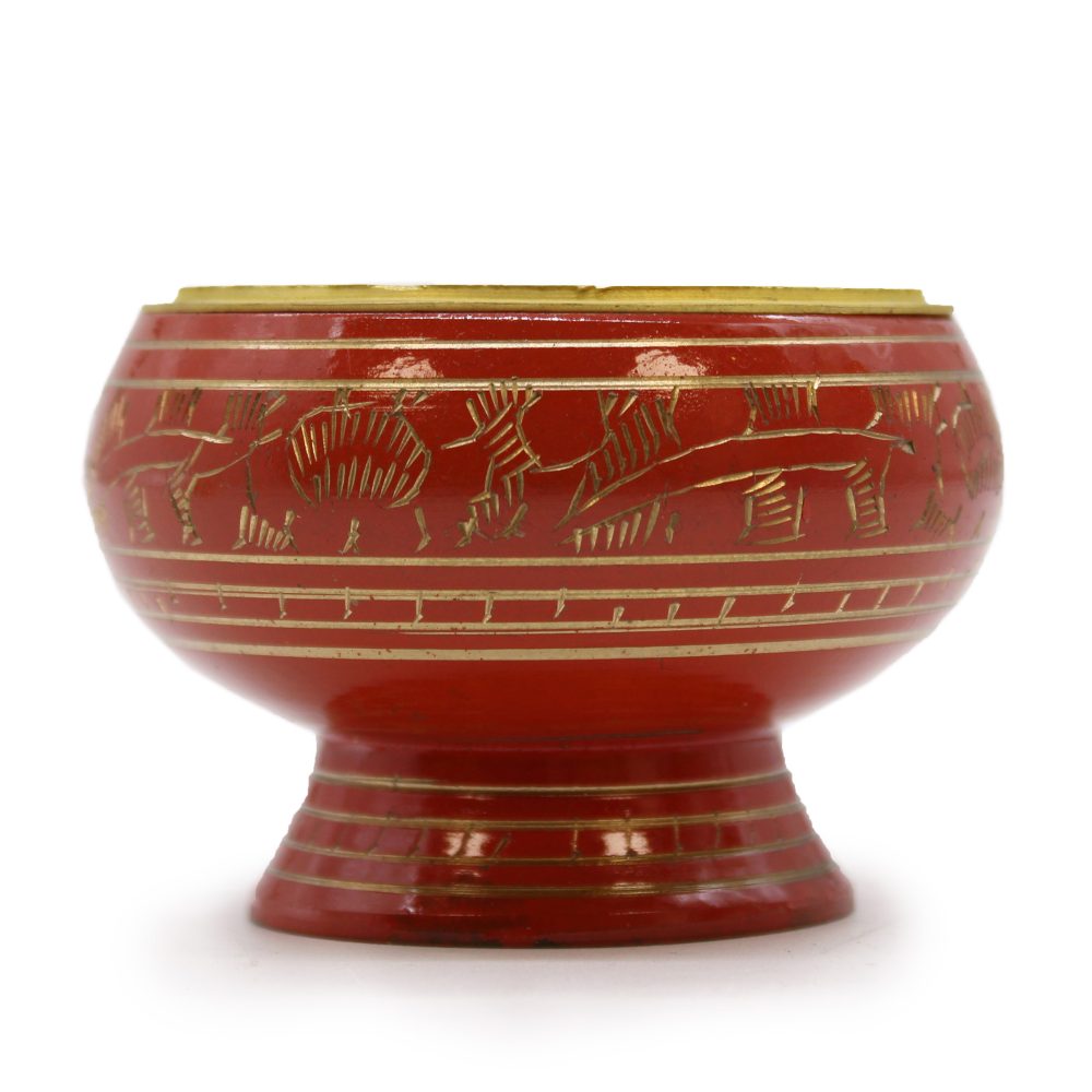 A red, decorative bowl stands on a flared pedestal base, featuring engraved gold animal motifs and horizontal lines, set against a plain, white background.