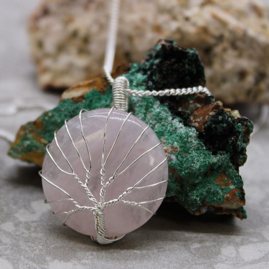 A pink, round gemstone pendant wrapped with silver wire in a tree design rests on a mottled green and brown stone. A silver chain extends from the pendant.