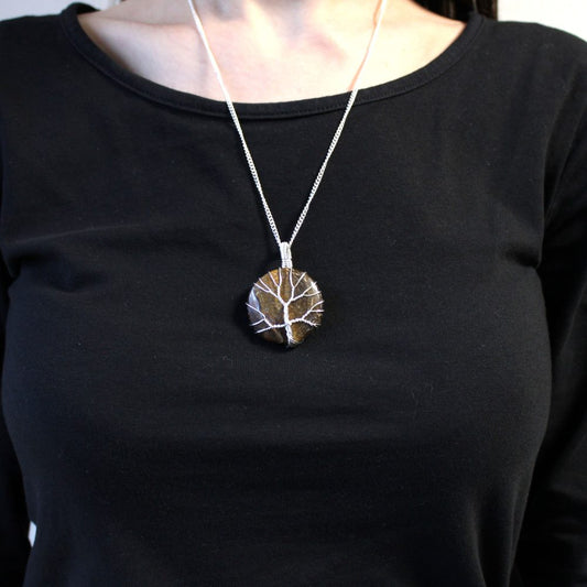 A pendant hangs on a silver chain around a person’s neck. The circular pendant has a brown stone and silver wire depicting a tree. The person wears a black long-sleeved shirt.