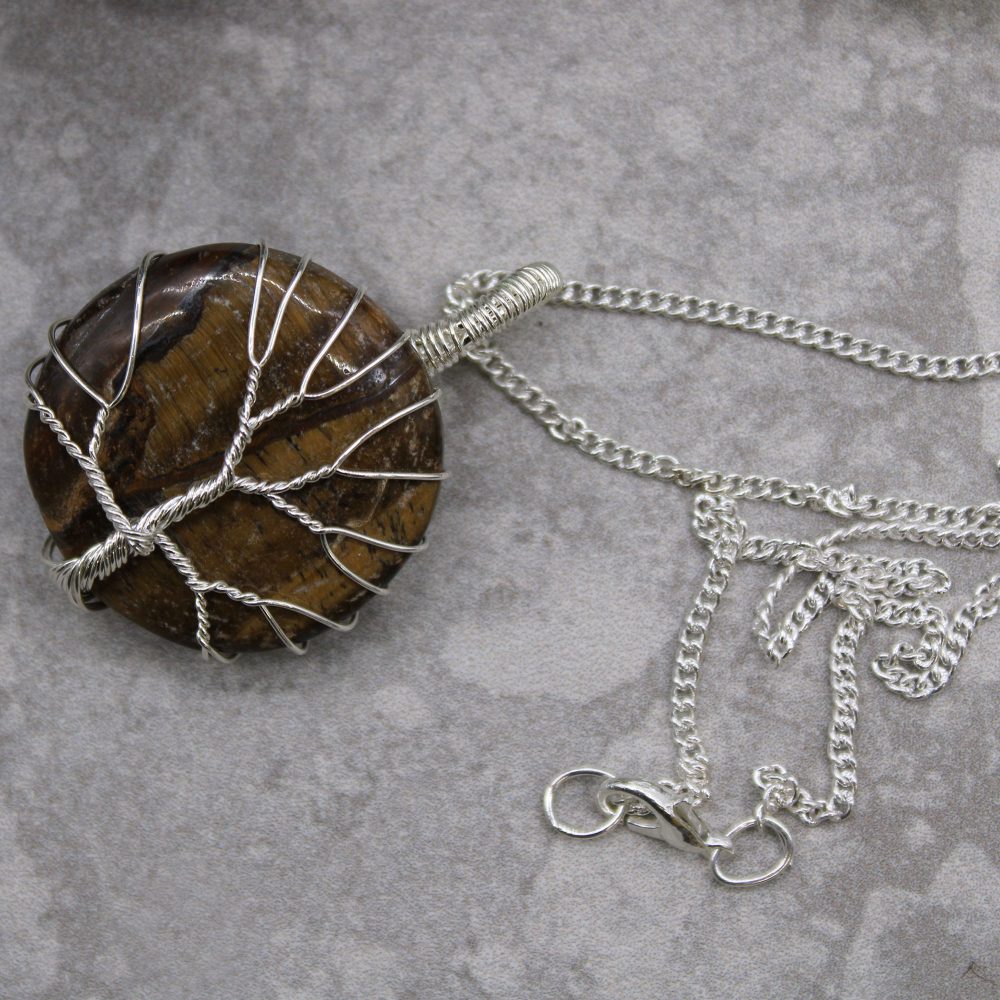A brown, disc-shaped stone pendant is wire-wrapped in a tree-of-life design. It lies on a gray surface, connected to a silver chain.